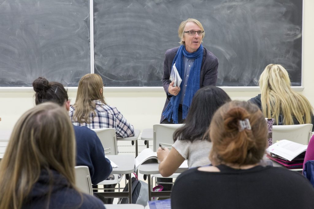 An instructor stands in front of a chalkboard in a classroom, engaging with students seated at their desks, who are listening and taking notes.
